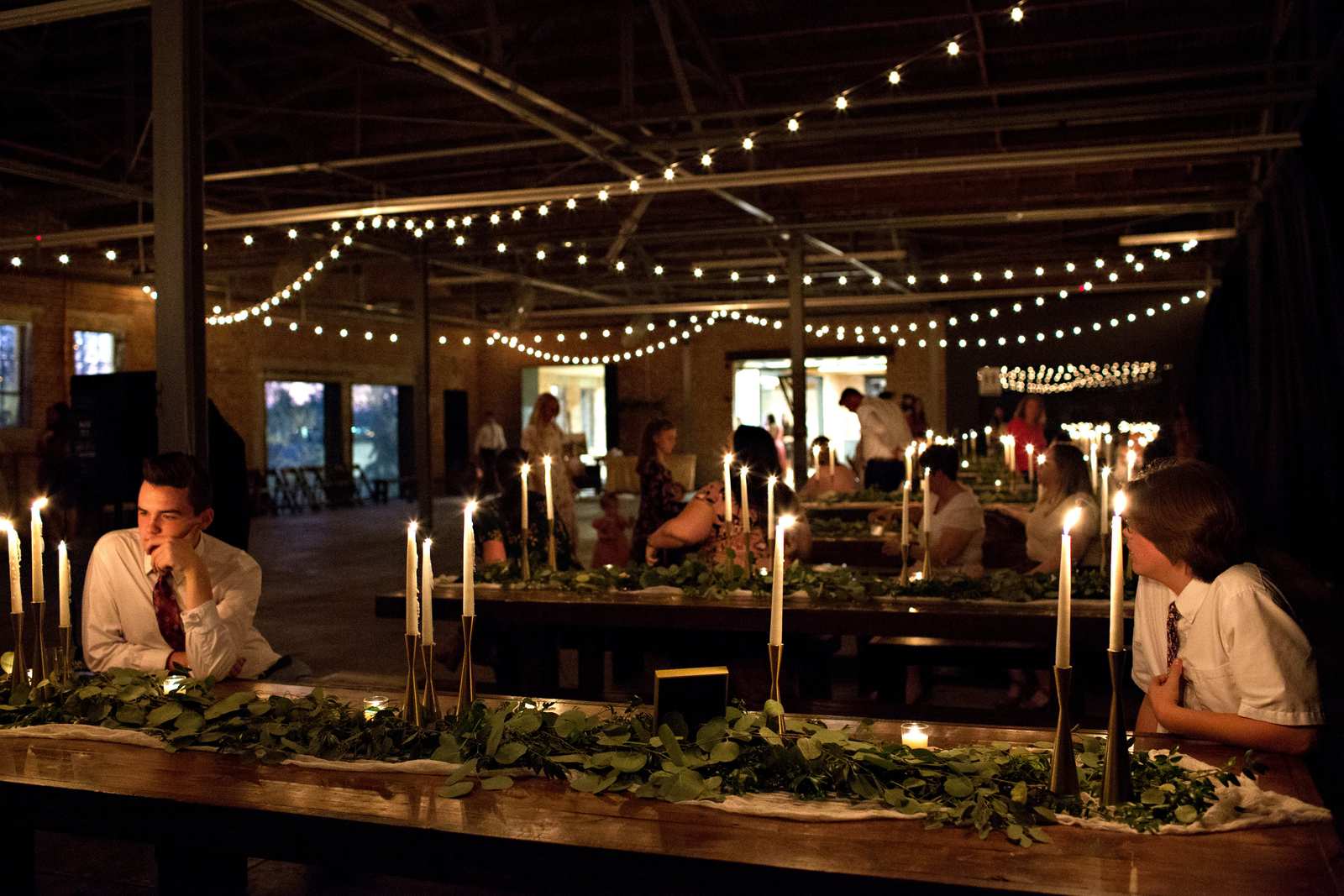 Guests dining at candlelit long tables dressed with greenery under the warehouse trusses