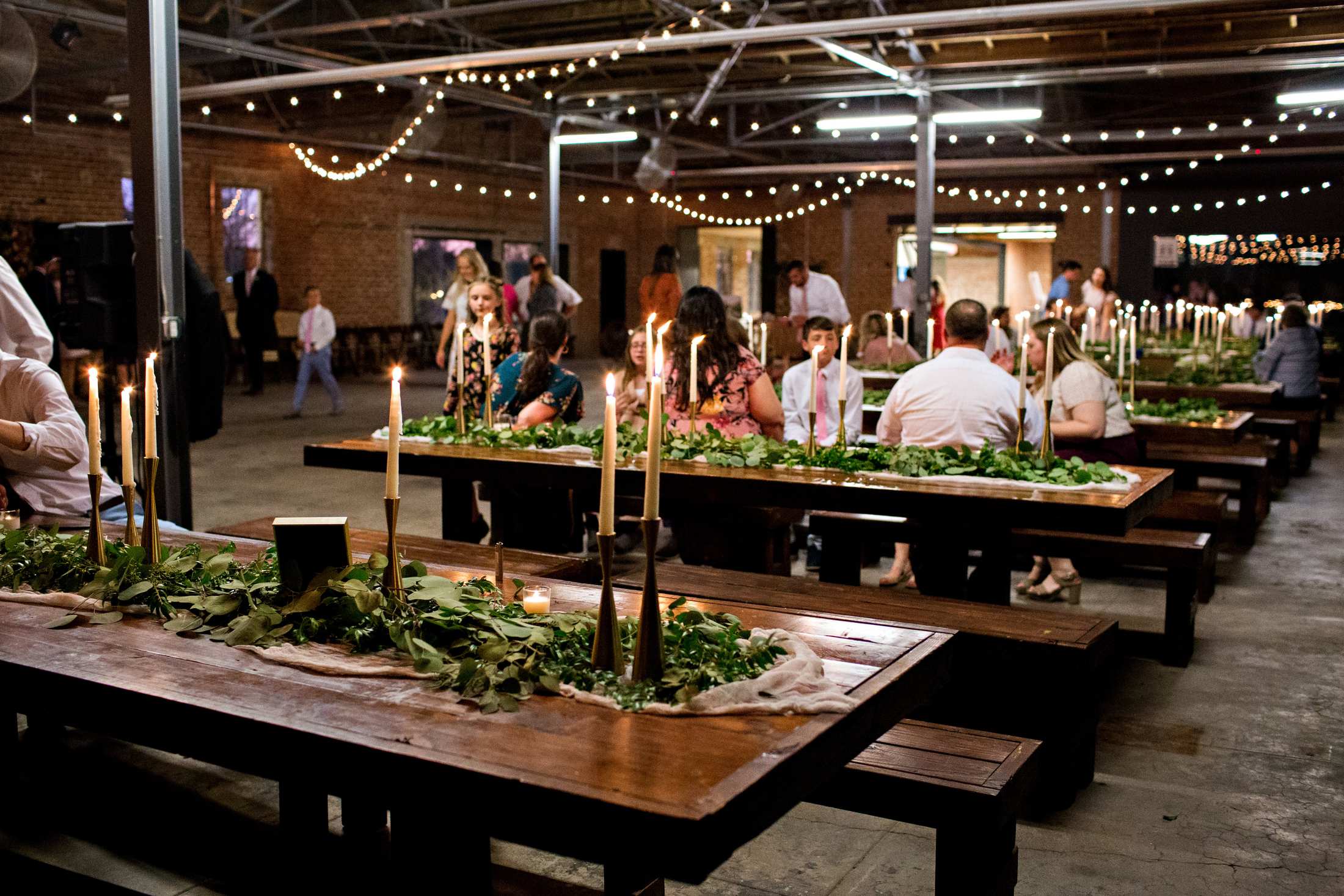 Long candlelit tables under warehouse string lights, guests seated at a wedding reception